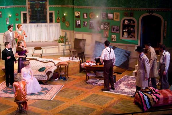 A theater scene with nine actors in a vintage living room set. Some wear elegant attire, others trench coats. Smoke rises from a small stove. The backdrop includes green walls, framed photos, and musical instruments. Rugs and furniture complete the setting.