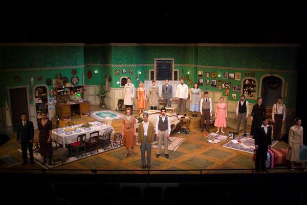 A stage production featuring actors in period costumes, set in a detailed room with green walls and vintage furniture. The scene includes tables, chairs, and various decor items, suggesting a mid-20th-century household atmosphere.