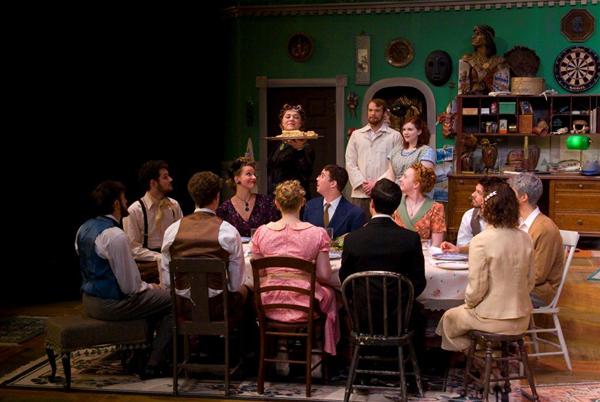 A group of people sit around a dining table in a theater setting, dressed in vintage clothing. A woman stands holding a platter. The background is decorated with various items, including a dartboard and sculptures. They appear to be engaged in a dinner scene.