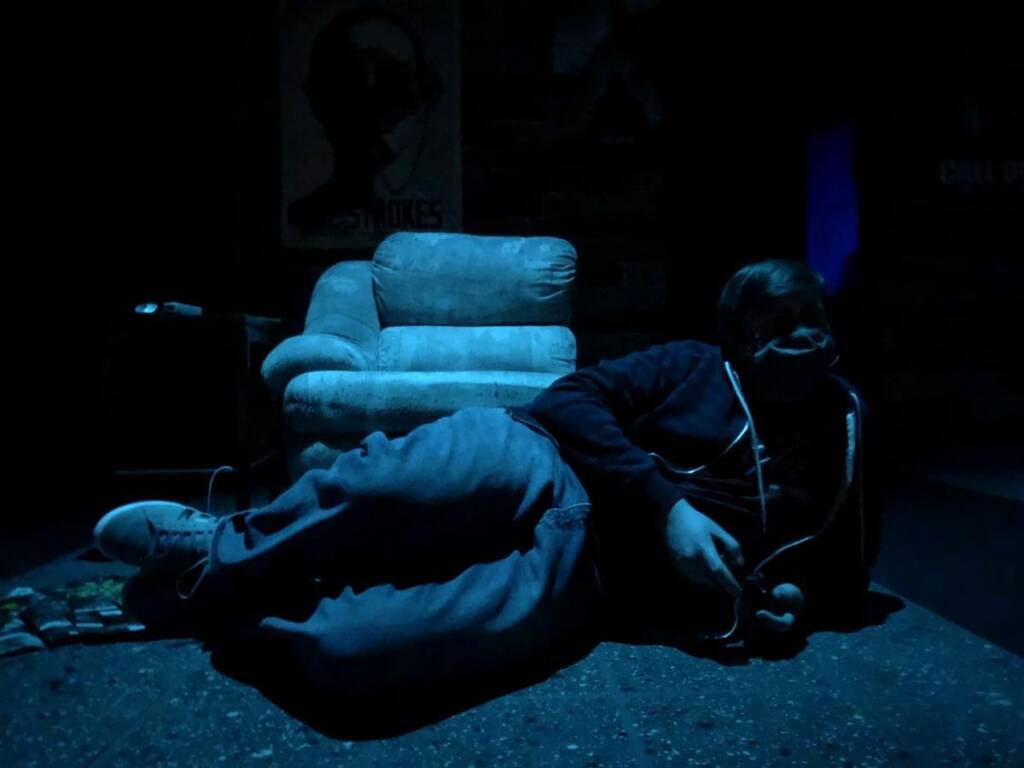 A person lies on the floor in a dimly lit room next to a recliner chair. They are wearing casual clothes and a mask, holding a remote control. A poster is visible on the wall in the background.