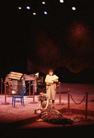 Three actors perform on stage. One lies on the floor covered in a patterned blanket, while another stands nearby looking at a third actor sitting on a chair. The set includes scattered furniture and a rope barrier with a backdrop resembling rock formations.