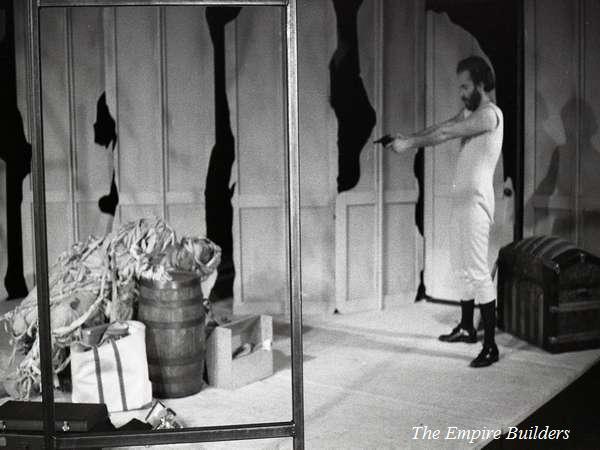 A man in a white outfit and boots points a gun in a theater set. The stage features barrels, wooden crates, and torn paper walls. The text "The Empire Builders" appears at the bottom right. The scene is monochrome.