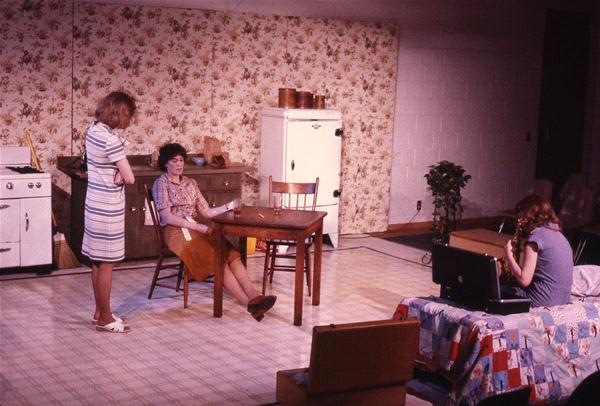 Three women are in a vintage kitchen setting. One sits at a table with her feet on a chair, another stands nearby, and the third is seated with a typewriter. The room has floral wallpaper, a stove, refrigerator, and quilt-covered furniture.