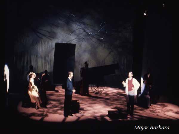 A stage scene from a theatrical production titled "Major Barbara." It features four actors in period costumes, with a large silhouette of a cannon in the background. The lighting creates dramatic shadows on the stage.