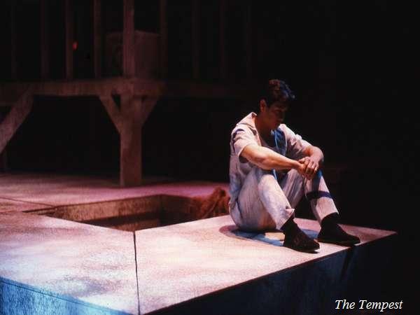 A person is sitting alone on a stage set, illuminated by a spotlight. They are wearing casual clothes and black shoes. The setting has a rustic, wooden structure and is labeled "The Tempest" in the bottom right corner.