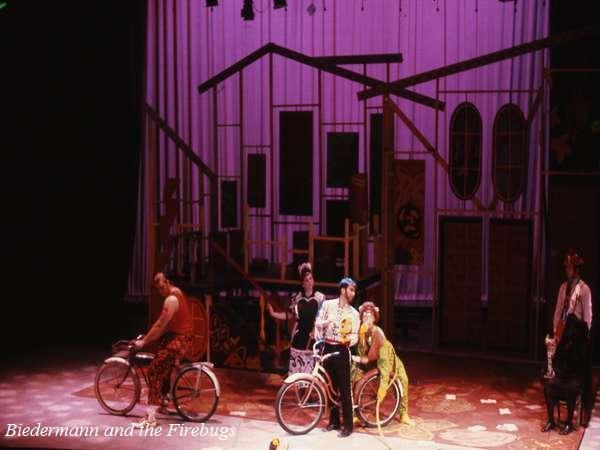 A whimsical stage scene from "Biedermann and the Firebugs" features actors performing with bicycles against a backdrop of geometric house shapes. The stage is lit with purple and pink lighting, creating a surreal atmosphere.