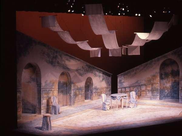 A theater stage set with two large, slanted walls featuring arched doorways. The scene includes a round table, a few chairs, and hanging white fabric strips. The backdrop is painted in earth tones with abstract designs.