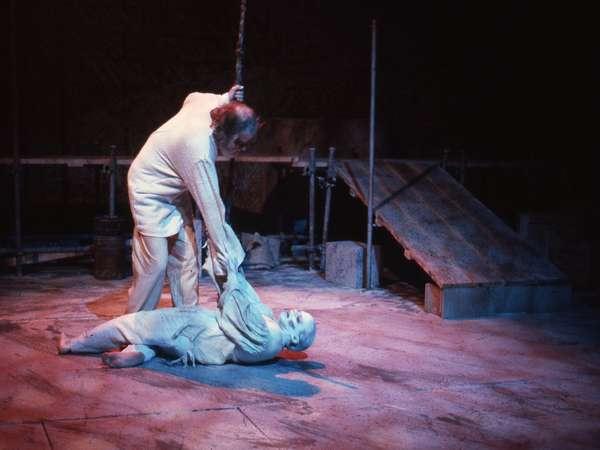 A stage scene features two actors. One actor stands, holding a rope, and is pulling a second actor lying on the floor. The floor is rugged, and the background includes a slanted platform, suggesting a dramatic setting.