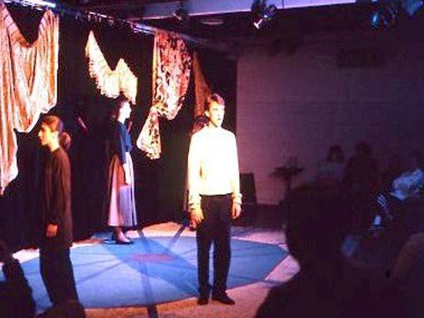 Three people are on a dimly lit stage with patterned fabric as a backdrop. One person in a white shirt stands in the center facing the audience, while the others stand on either side. The audience is visible in the shadows.