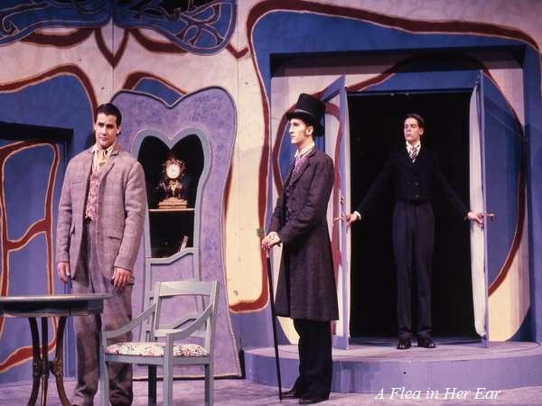 Three actors perform a scene on stage with a colorful, abstract backdrop. One actor stands in the foreground wearing a light gray suit, while the others, dressed in darker suits, stand in the background near a central doorway.