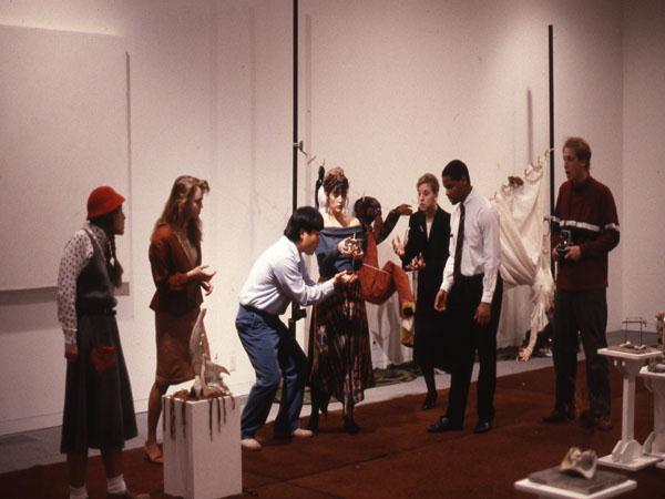 A group of seven people in a gallery setting, some holding props and interacting with art pieces. The scene suggests a blend of performance and art interpretation, with a casual atmosphere.