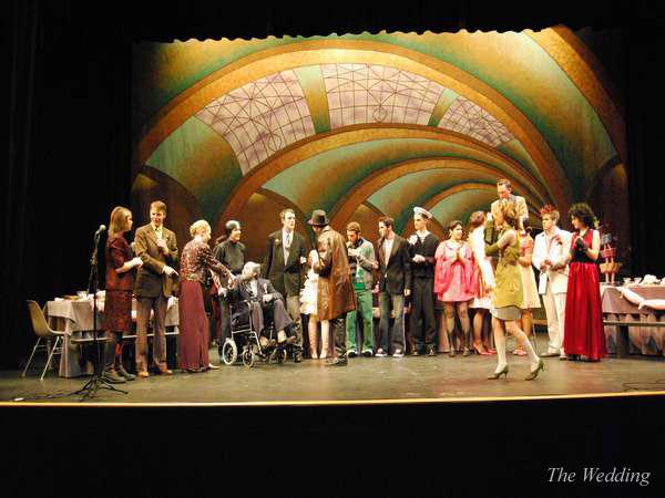 A group of people on stage performing "The Wedding." The scene features guests, one in a wheelchair. The backdrop has an ornate, arched design. The actors wear various costumes, including formal and vintage outfits. A table is set on the left.