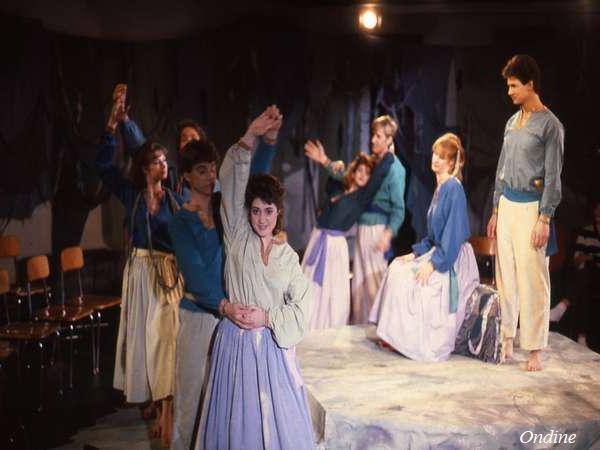 A group of people in a theatrical performance, dressed in blue tops and long skirts or pants, are on stage. One pair is dancing in the foreground while others sit or stand, watching. The backdrop is dark, creating a dramatic atmosphere.