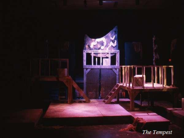 A dimly lit theater stage with wooden platforms, stairs, and a central backdrop shaped like a wave. The setup suggests a scene from "The Tempest.