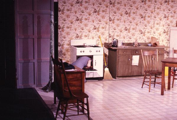 A person is partially inside an open oven in a vintage kitchen with floral wallpaper. Nearby are a wooden chair, table, broom, and cabinet. The scene has a retro, domestic setting with muted colors.