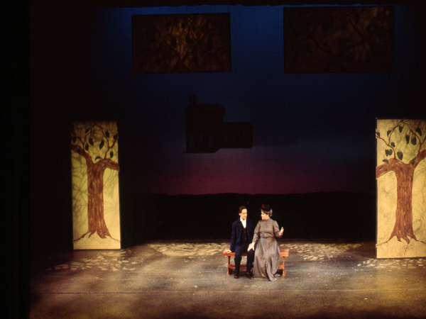 A theatrical stage with two actors. A man in a suit sits on a bench while a woman in a long dress stands next to him. The backdrop features a gradient sky and tree illustrations. The lighting casts soft shadows on the floor.