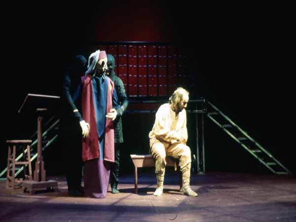 A dimly lit stage scene depicts a seated person in light-colored clothing, with another standing person behind them in dark clothing and a colorful cape. A wooden lectern is nearby on the left, and stairs in the background lead up on both sides.