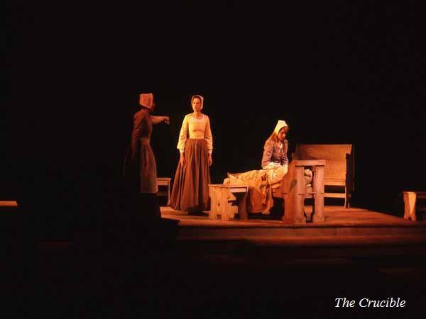 Three actors in period costumes perform a scene on stage. One stands pointing, another sits on a bench, and a third stands confidently. The set includes wooden benches. The stage is dimly lit. Text reads "The Crucible.