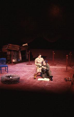 Two actors perform on a dimly lit stage. A woman sits on a chair while a man sits on the floor with an open book. The backdrop is dark with a few props, including a blue chair and some ropes.