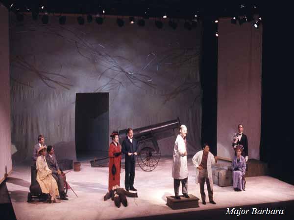 A theater stage set for a play with eight actors in period costumes. In the background, a cannon is visible with a painted sky backdrop. The scene includes some actors standing and others sitting, with various props around. "Major Barbara" is written in the corner.