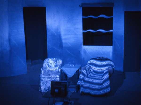 Two patterned armchairs face a small TV in a dimly lit room with blue lighting. A small table with glasses is between the chairs. A window with wavy lines is visible on the wall.