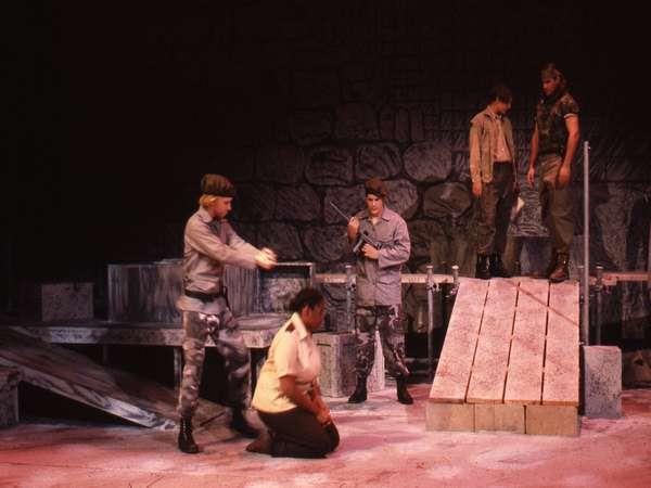 A group of actors performs on a stage with a dramatic setting. One actor kneels while another gestures emphatically. Two actors stand on a raised platform holding rifles, dressed in military-style outfits. The scene has a tense, military theme.