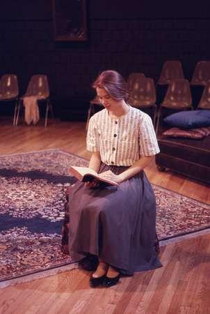 A person in vintage clothing sits on a wooden floor, reading a book. They wear a striped blouse and a long skirt. In the background, empty chairs and a couch are visible. The setting has a warm, intimate ambiance.