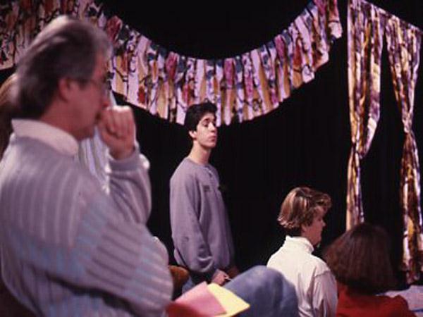 A group of people, three seated and one standing, are in a room with colorful, abstract fabric banners hanging in the background. The atmosphere seems contemplative and artistic.