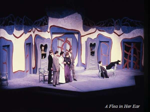 Stage set for a production of "A Flea in Her Ear" featuring five actors in period costumes on a colorful, surreal set with exaggerated doorways and windows. A person sits on a chair while others stand grouped together.