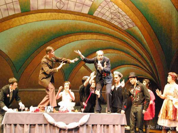 Performers on stage act out a dynamic scene with lively gestures. The colorful set features an arched ceiling design. Participants stand atop a long table covered with a cloth, engaging energetically with one another.