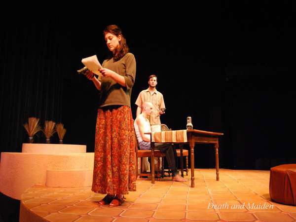 A woman stands reading a script on stage, wearing a long skirt and blouse. Behind her, two men sit at a wooden table with papers and a glass. The stage is dimly lit, with props including a jug and vases. The text "Death and Maiden" is visible.