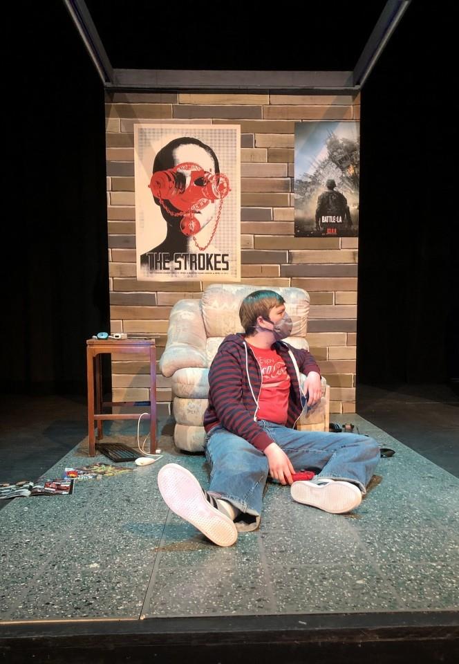 Man sitting on the floor in front of an armchair, looking to the side. Behind him are posters on a brick wall, including one of a band and a movie. He's wearing a striped hoodie and jeans, and the floor has scattered items. The setting resembles a room or stage set.