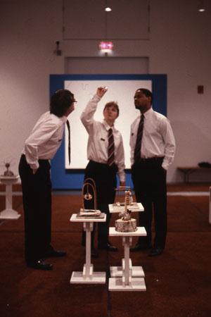 Three people in white shirts and ties examine an art exhibit with small sculptures on pedestals. One person gestures toward the ceiling. The room has a large, blue-framed screen in the background and is carpeted in brown.