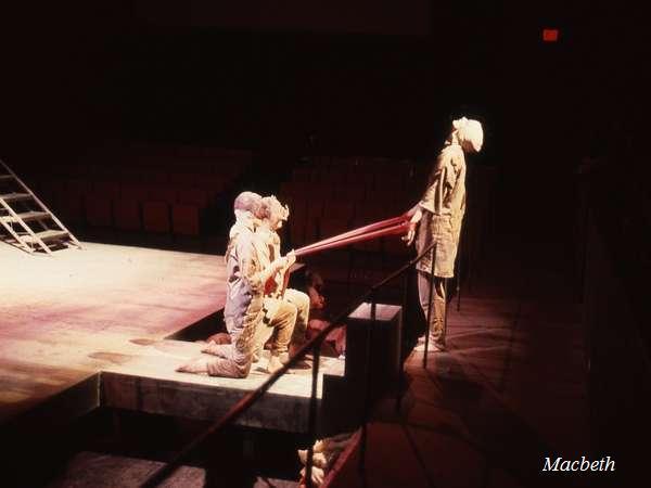 Three actors in medieval-style costumes perform on a dimly lit stage. Two kneel, holding long objects resembling swords or staves, directed toward a standing figure. The background is dark, with empty theater seats visible. Text reads "Macbeth.