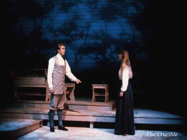 Two actors perform on stage in a scene from "The Crucible." The man wears historical clothing with a vest and boots, gesturing towards the woman who is in a long skirt and blouse. Wooden benches and a blue-lit background set the atmosphere.