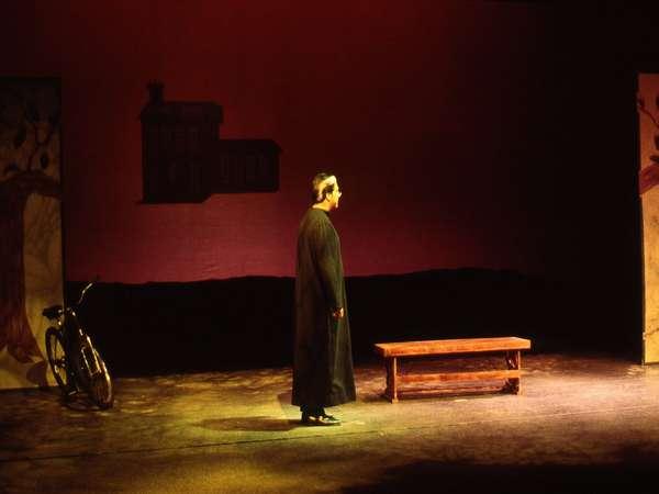 A person in a long, dark robe stands on a dimly lit stage. There is a bicycle leaning against the backdrop and a wooden bench nearby. The walls are painted with trees and a house silhouette. The lighting is warm and dramatic.