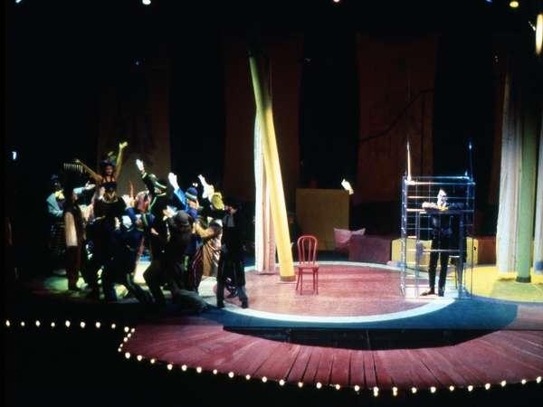 A circus stage with performers reaching towards a figure on the right, who is inside a tall cage. A single red chair sits empty on the stage. The scene is lit dramatically with spotlights, creating an atmosphere of anticipation.