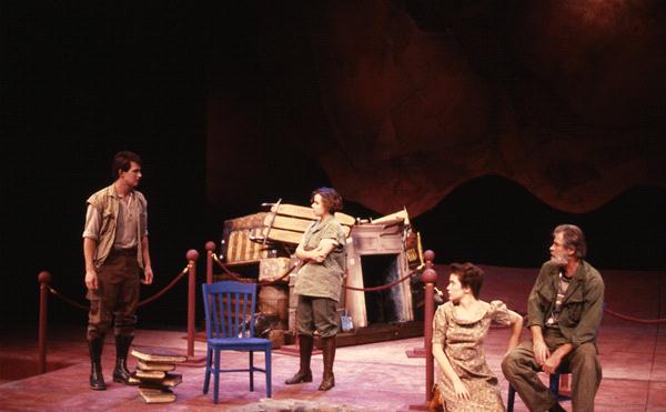 Four actors on stage during a play. One stands on the left beside a stack of books and a blue chair, engaging with the others to the right. The background features a piano and draped ropes, suggesting a theatrical set.