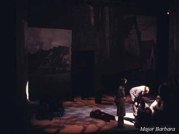 Dimly lit stage scene from "Major Barbara" with a group of actors huddled near the bottom right. Shadowy projections of buildings are in the background, and a beam of light highlights part of the scene.