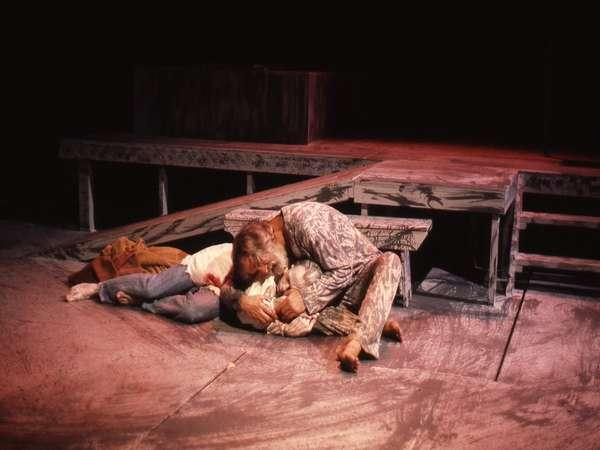Two actors on stage in an intense scene. One person leans over the other, who is lying on their back. The stage is set with rustic, wooden platforms, creating a dramatic atmosphere.