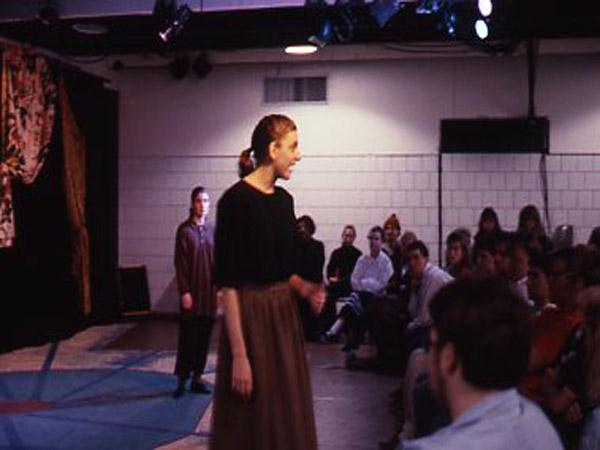 A person on stage passionately speaks to an audience seated in a small theater. Another person stands in the background. The setting is intimate, with stage lights illuminating the scene.
