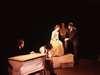 A theatrical scene with four actors on stage. Two people are sitting at separate desks, one engaged in writing. A man and a woman in costumes are standing and interacting in the center. Dim lighting creates a dramatic atmosphere.