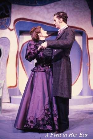A woman and a man in period costumes stand closely together, gazing at each other on stage. The woman wears a purple dress, and the man is in a dark suit. The backdrop features abstract shapes. Text reads "A Flea in Her Ear.