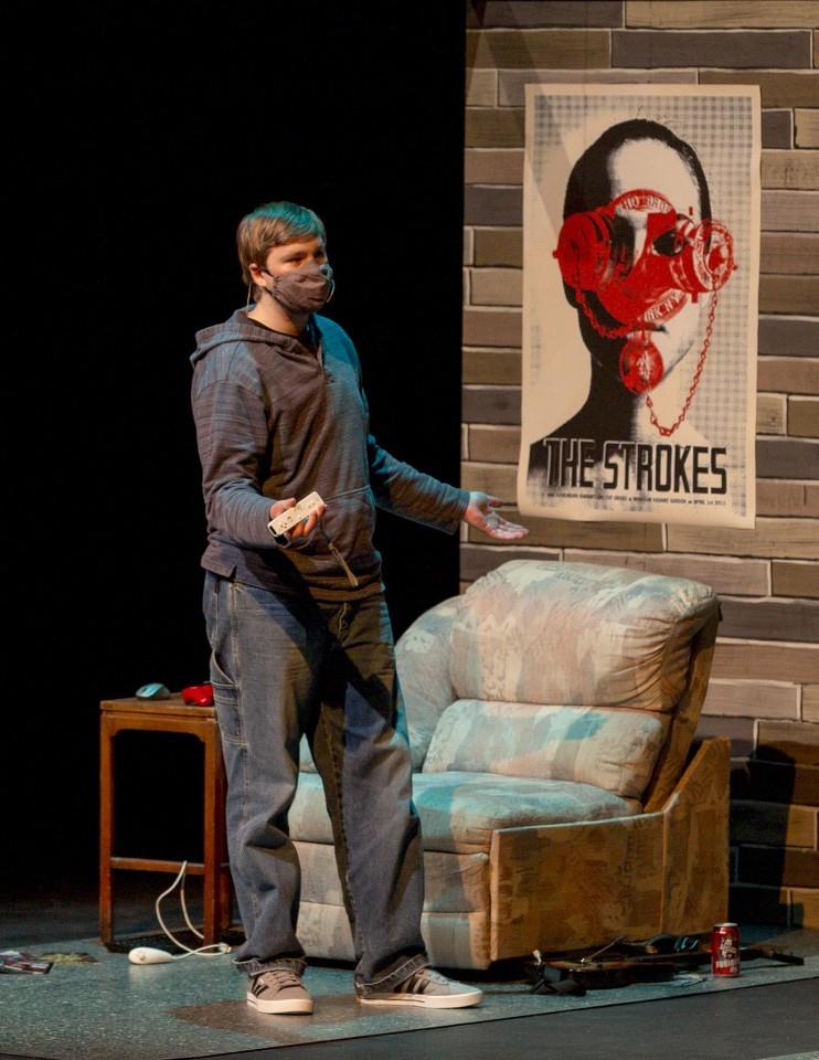 A person stands on stage wearing a mask, holding a video game controller, near a chair and small table. A poster of The Strokes is on the wall behind them. The scene suggests a casual setting.