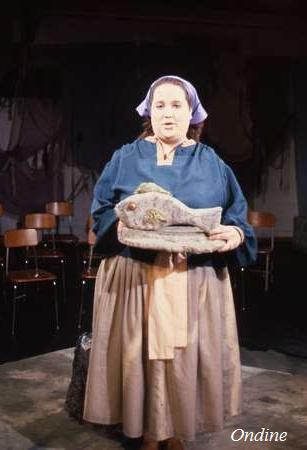 A person wearing a blue top, brown skirt, and purple headscarf holds a decorative fish sculpture. They're standing in a dimly lit room with several wooden chairs in the background. The word "Ondine" is seen in the bottom right corner.