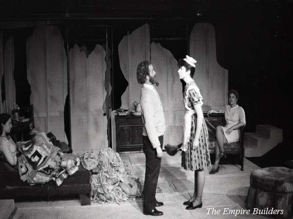 A black and white theater scene shows four people on stage. A man and woman stand facing each other in the center. Another woman sits on a couch, while a fourth person sits in the background. The set includes abstract, painterly walls. Text reads "The Empire Builders.