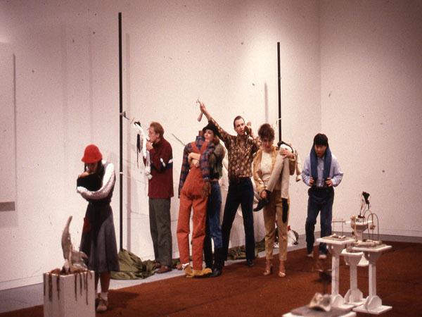 A diverse group of people stands in an art gallery. One person is wrapped in a coat, others appear to be posing creatively with sculptures. The gallery has various abstract art pieces and sculptures on display.