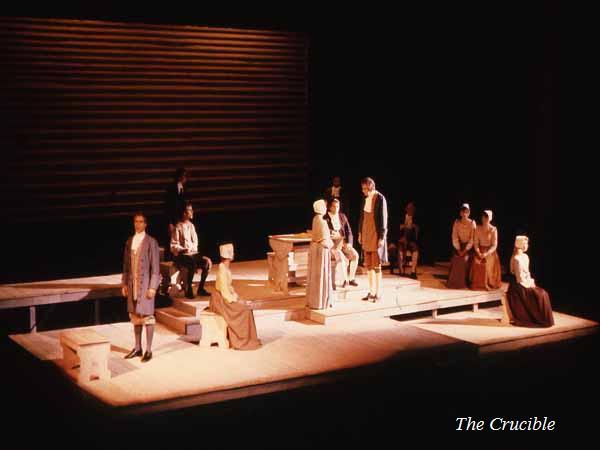 Stage production of "The Crucible" with actors in period costumes on a minimalist set. The scene includes benches and a table, with dim, focused lighting creating a dramatic atmosphere.
