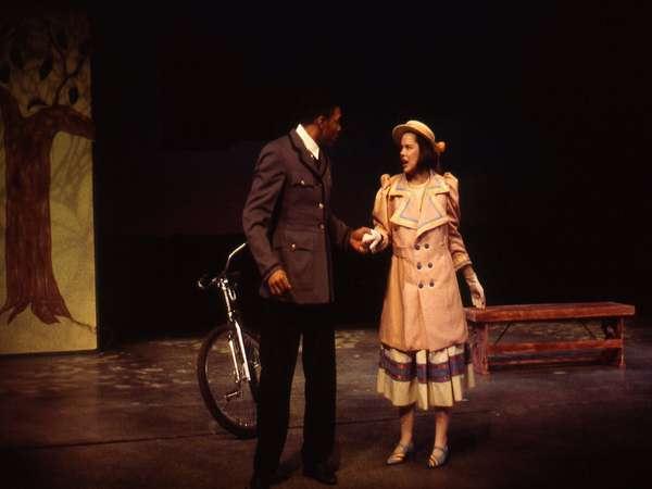 A man and a woman in vintage attire are standing on a dimly lit stage. The man wears a dark suit, and the woman is in a beige dress and hat. A bicycle is seen behind them, next to a painted tree and a bench.