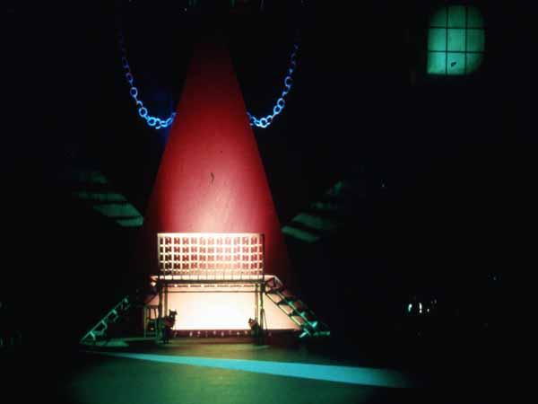 A dimly lit stage with a large red cone structure at the center. Chains hang from the top of the cone. A metallic frame with stairs on both sides is positioned in front, illuminated by a spotlight.